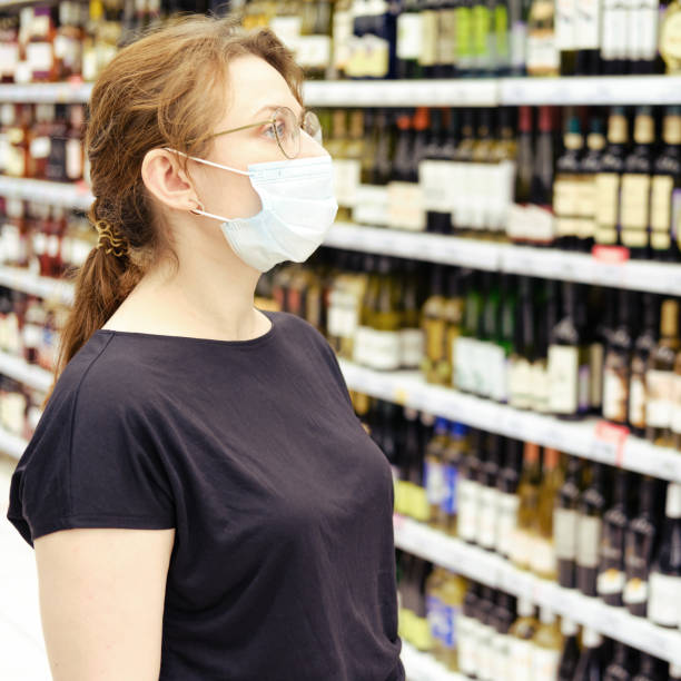 une femme adulte dans un masque médical se tient dans un département avec le vin dans un hypermarché. vendre de l’alcool dans un supermarché. être dans des lieux publics pendant l’épidémie du coronavirus - eastern european caucasian one person alcoholism photos et images de collection