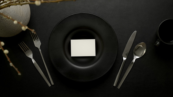 Top view of dark luxury dinning table setting with mock-up place cacd on black ceramic plate, silverware and decoration