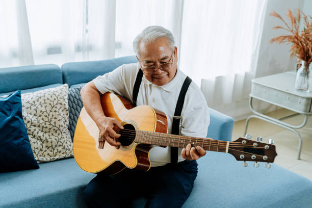 hombre mayor asiático sentado en el sofá y tocando la guitarra acústica. feliz sonrisa anciano cantando y disfrutando con la guitarra. disfrutar de la vida de jubilación en casa. - guitar playing music human face fotografías e imágenes de stock