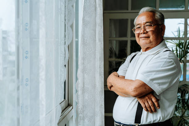 Confident ethnic man standing near window at home Positive plump senior Asian male smiling for camera and crossing arms while resting near window in cozy room at home asian culture stock pictures, royalty-free photos & images