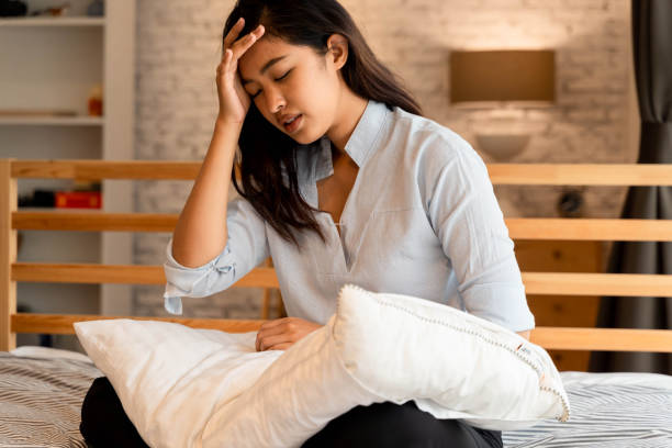Young beautiful Asian woman having a headache while lying on bed, feeling sad and stressed at home. Disappointed and desperate girl crying stock photo