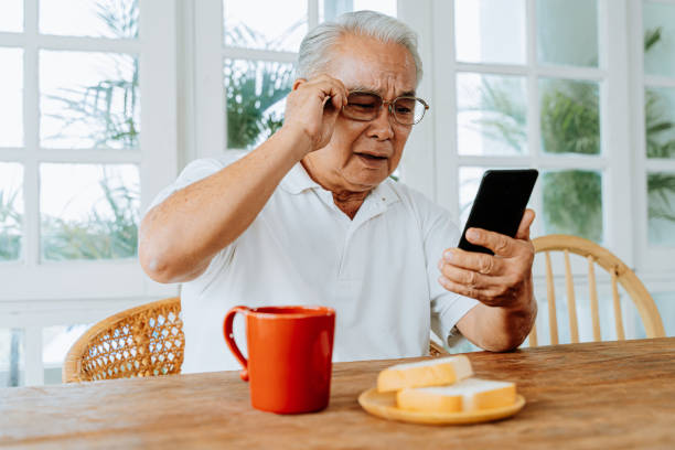istock Elderly Asian male having eyesight problems and wearing eyeglasses while gazing and reading message on smartphone during breakfast at home. Old man having eye blurred vision indoors 1253588784