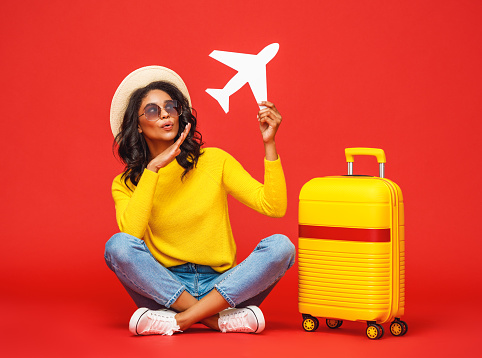 Full body ethnic female in hat and sunglasses sitting cross legged near suitcase and playing with toy airplane against red backdrop