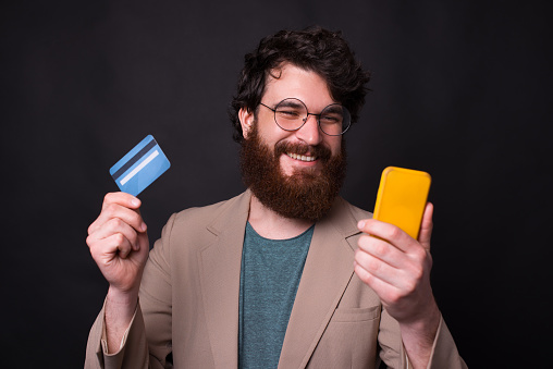 Cheerful young man with beard in casual using smartphone and credit card.