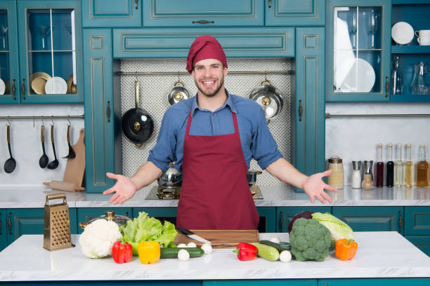 istock Man smile in chef hat in kitchen. Happy cook at table. Vegetables and tools ready for cooking dishes. Food preparation and cooking recipes. Vegetarian menu and healthy diet 1253618769