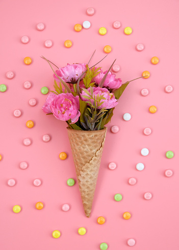 Waffle cone with flowers and colorful candy on pink background. Decorations for birthday, anniversary, wedding. Minimal decorative holiday concept.