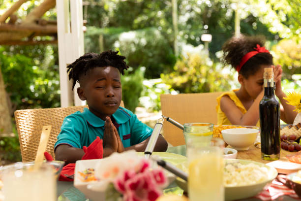 Family eating together at table Front view of a young African American brother and sister sitting at a table with eyes closed and hands in prayer, saying grace before having a meal with their family outside on a patio in the sun saying grace stock pictures, royalty-free photos & images