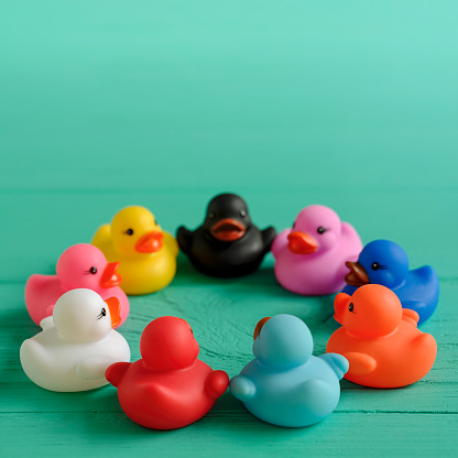 A group of rubber ducks of many different colors sit in a circle opposite each other as if discussing and talking about their differences but being together peacefully on an old wooden turquoise-colored grained table background. Concept image representing discussion, negotiation, peace, living together, race, gender, ethnicity, living in harmony, politics, diversity, LGBTQI, etc.