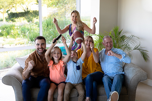 Caucasian multi-generation family in the living room at home, sitting on a couch together, raising hands and cheering while watching TV, the grandson holding an American football