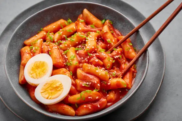 Tteokbokki with eggs in gray bowl on concrete table top. Tteok-bokki is a korean cuisine dish with rice cakes. Asian food.