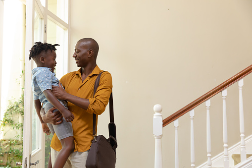 Side view of an African American man arriving home, wearing a shoulder bag and carrying his young son, standing in the hallway and smiling at each other