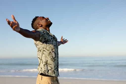 A happy, attractive African American man enjoying free time on beach on a sunny day, wearing a Hawaiian shirt, sun shining on his face with his arms outstretched. Relaxing summer vacation.