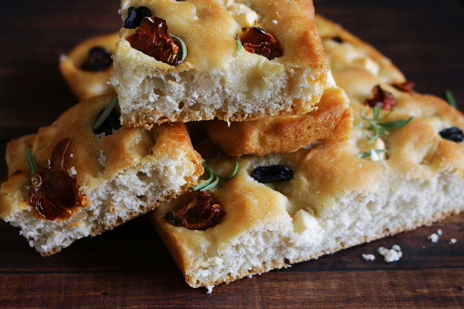 Stock photo showing square slices of flat Italian focaccia bread, which is homemade and has just been taken out of the oven, being pictured with a garnish of rosemary leaves. This traditional flat bread features green olives, sun-dried tomatoes and feta cheese, as well as olive oil, herbs and sea salt.
