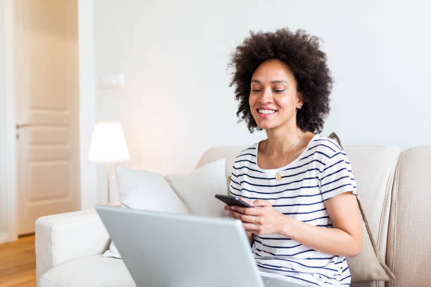 istock Happy young black woman using mobile phone while sitting a couch at home with laptop computer. Smiling woman sitting on sofa relaxing while browsing online shopping website. 1253635107