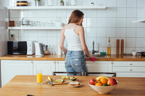 back view of young woman preparing breakfast in modern kitchen near table with fresh fruits