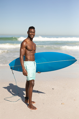 A portrait of a happy, attractive African American man enjoying free time on beach on a sunny day, smiling, having fun, standing with his surfboard, sun shining on him.