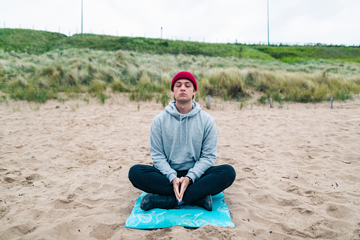 Front view of a caucasian man dressed warmly doing yoga and meditation at a beach early in the morning. He is doing the easy pose and is in the North East of England.