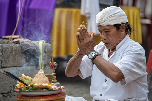 Pemangku (Balinese priest) praying at the yard of a house, during a celebration for the third month baby anniversary. He clasps his hands reciting prayers as the incense burns. Pemankus are low chaste priests, who serve in family ceremonies, temple maintenance and regular offerings. Most have daily jobs, and their own families.