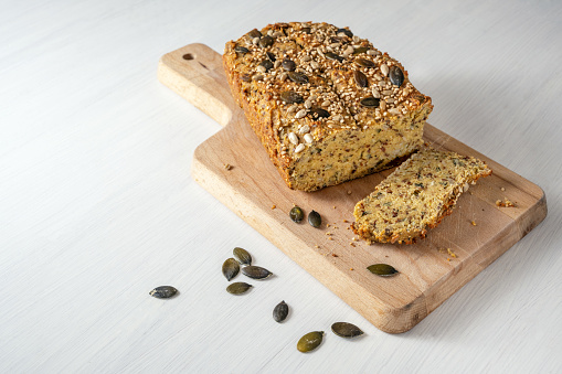 Protein bread from quark, oat bran, lupine flour, almond, pumpkin seeds and other healthy ingredients, baking for low carb or ketogenic diet, wooden cutting board on a white table, copy space, selected focus, narrow depth of field