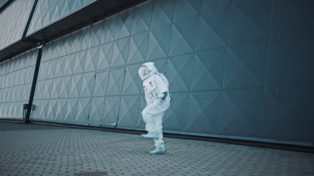 Handsome Man in Spacesuit is Dancing Next to Metal Wall. Astronaut is Happy and Makes Creative Robotic Moves. Successful Spaceman in White Futuristic Suit with Technological Panel on His Hand.
