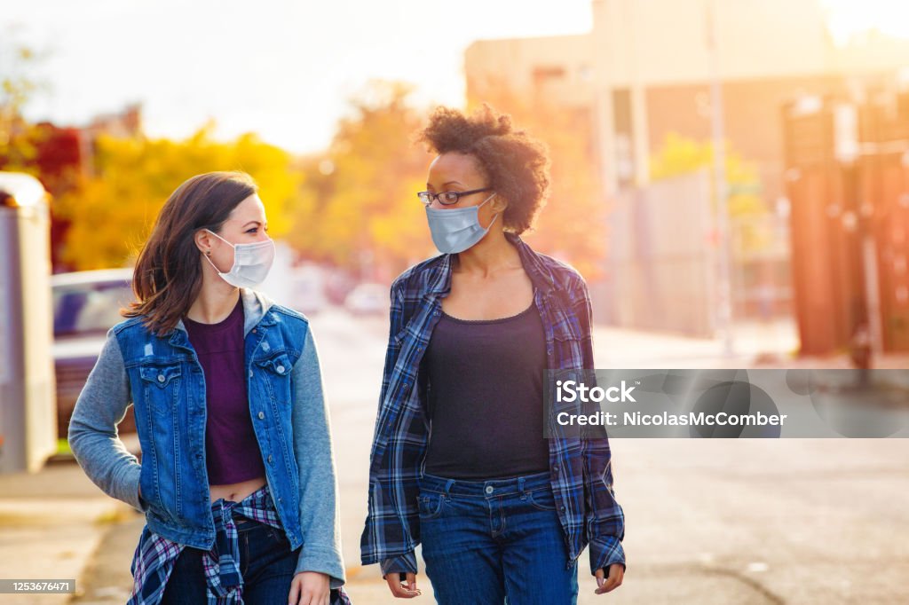 Two female friends walking down a Brooklyn alley wearing face masks Two female friends walking down a Brooklyn alley wearing face masks on a sunny Autumn day, having a quiet conversation. Protective Face Mask Stock Photo