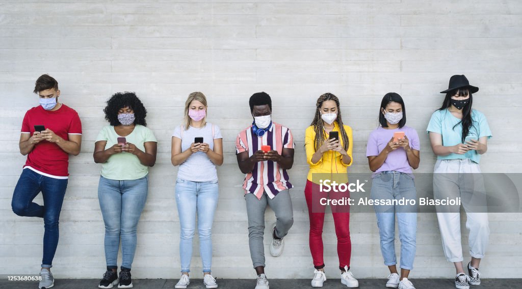Young people wearing face mask using mobile smartphone outdoor - Multiracial friends having fun with new technology social media app during corona virus outbreak - Youth millennial lifestyle concept Social Media Stock Photo