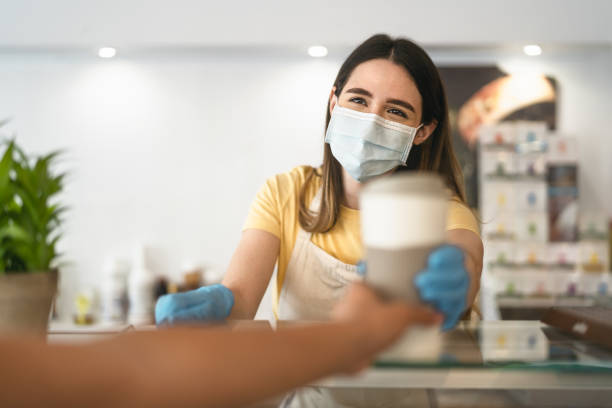 bar owner working only with take away orders during corona virus outbreak - young woman worker wearing face surgical mask giving coffee to customer - healthcare and drinks concept - foods and drinks clothing garment household equipment imagens e fotografias de stock