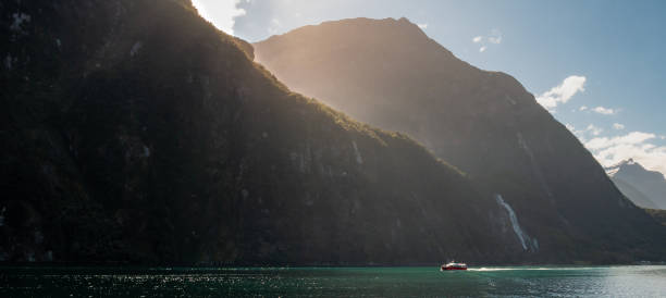 tiro panorâmico de barco navegando através de fiord em um dia ensolarado com raios solares criando aura dourada ao redor da borda da montanha - hiking coastline waters edge sunny - fotografias e filmes do acervo