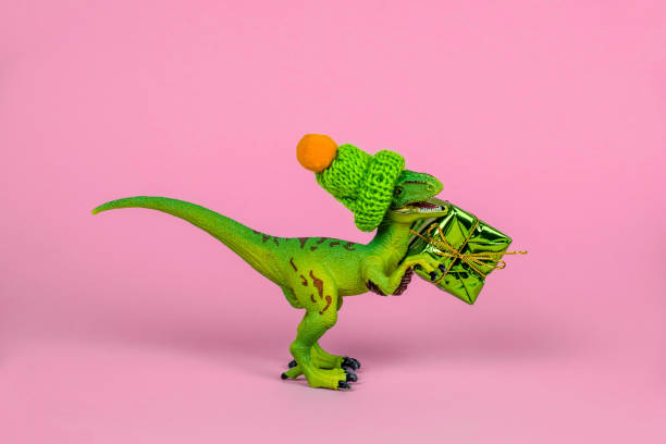 istock cute green plastic toy dinosaur wearing knitted hat and holding present box 1253691704
