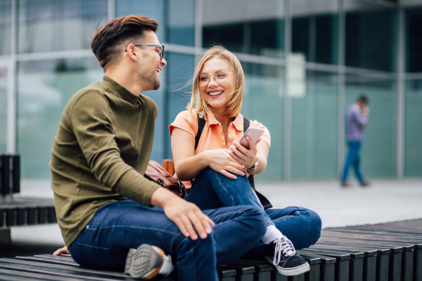 deux étudiants souriants s’asseyant sur un banc de stationnement - smart casual outdoors friendship happiness photos et images de collection