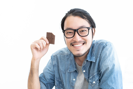 Happy young bearded hipster Asian man in eyeglasses eating a chocolate bar isolated over white background.