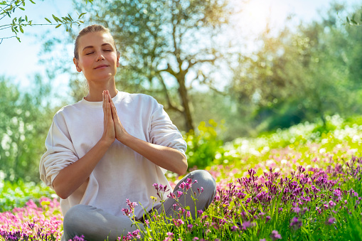 Beautiful woman doing yoga exercise outdoors, nice female in lotus pose meditating with closed eyes in beautiful blooming spring garden, healthy lifestyle, relaxation and wellness concept