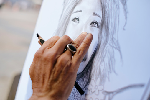 Mature Man Working As Street Artist Painter, Drawing Portrait Of Tourist On Canvas In Town Square, Madrid, Spain.
