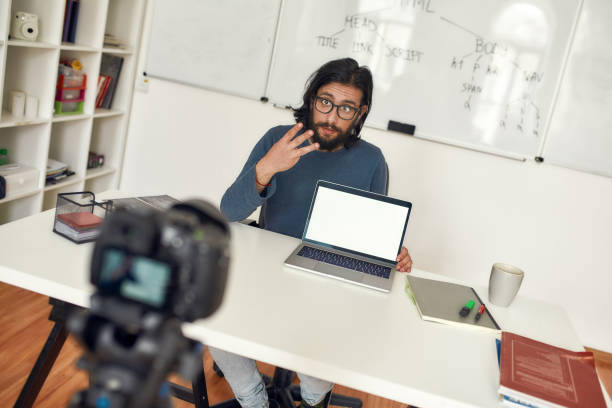 istock Young male teacher sitting at his workplace and explaining new theme while recording video blog on professional digital equipment. Coding class online 1253715892