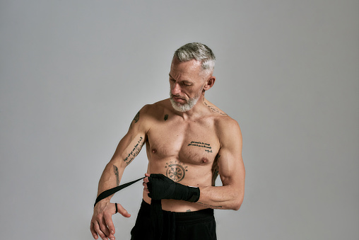 Half naked middle aged athletic man, kickboxer wrapping hands for Muay Thai, Boxing or Kickboxing, standing in studio over grey background. Sport, healthy lifestyle concept. Front view