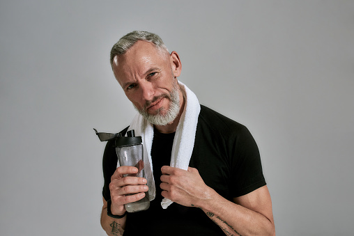Middle aged muscular man in black t shirt looking at camera holding bottle of water and towel around his neck, posing in studio over grey background. Sport, healthy lifestyle concept. Horizontal shot