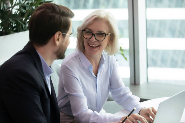Elderly businesswoman sitting next to subordinate Smiling elderly businesswoman wearing glasses, work at laptop, sit next to subordinate man and look at him, successful female boss in modern office subordination stock pictures, royalty-free photos & images