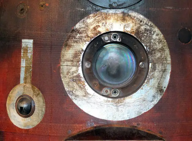a close up of a round porthole on a used russian space re-entry vehicle with burned red and black steel panel