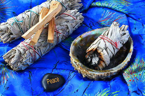 And image of white sage bundles in an abalone shell on a bright blue silk background.