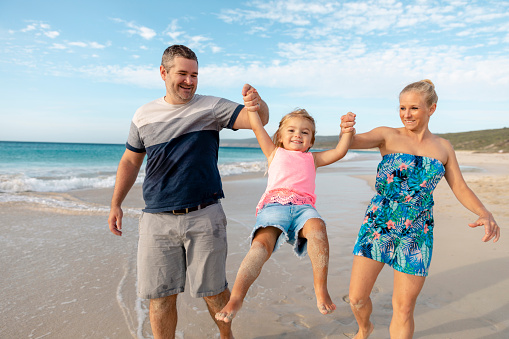 A shot of a caucasian family visiting the beach on a sunny day in Perth, Australia. Parents are swinging their daughter in the air.