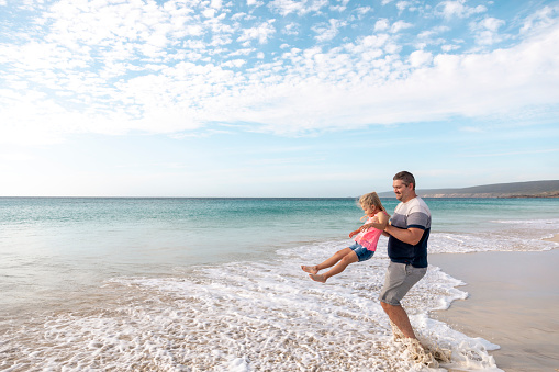 A shot of a caucasian family visiting the beach on a sunny day in Perth, Australia. A father is holding his young girl in the air, she is having fun.