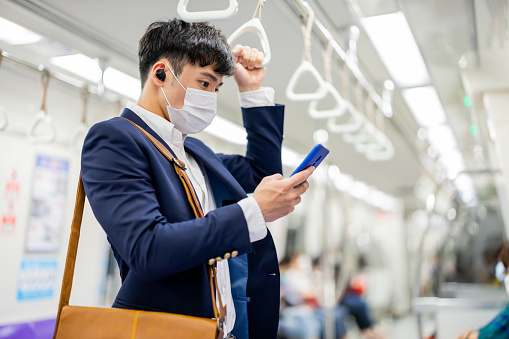 Asian business man wear surgical mask face protection and use smart phone with ear bud while commuting in the metro or train