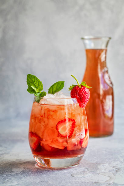 A red refreshing drink with ice, decorated with mint leafs and a strawberry. With a bottle of red drink in the background. A red drink with ice and sliced strawberries, garnished with mint and a fresh strawberry., and a carafe of the same liquid in a the background. The drink is on a gray background with copy space. Mocktail cordial strawberry drink. strawberry crush stock pictures, royalty-free photos & images