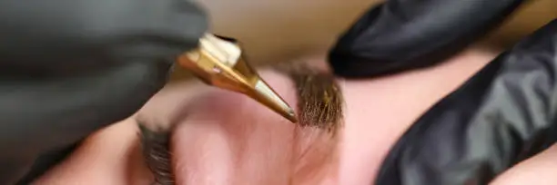 Close-up of professional makeup artist using golden tool and applying colour on eyebrow shape. Macro shot of specialists hands in protective gloves. Beauty and permanent procedure concept