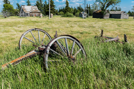 Vintage wagon wheel in front of old, abandoned prairie farmhouse with trees, grass and blue sky in Saskatchewan, Canada
