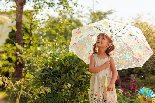 Little girl with an umbrella looks at the sky. Beautiful girl in garden under the summer rain. Vacation in village.