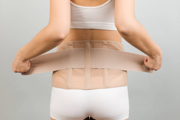 back view of pregnant woman in underwear dressing orthopedic corset to make the backache go away at gray background with copy space. close up of orthopedic abdominal support belt concept - bustiers imagens e fotografias de stock