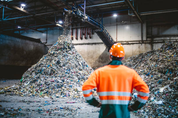 Worker Observing Processing of Waste at Recycling Facility Rear view of young male worker in helmet, pollution mask, and reflective clothing observing waste falling from conveyor belt onto pile at facility. rubbish heap stock pictures, royalty-free photos & images