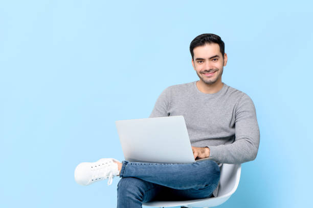 istock Portrait of smiling young handsome Caucasian man looking at camera while sitting using laptop in isolated studio blue background 1253818238
