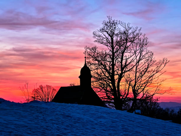 Silhouette of a chapel behind a snow covered hill with a dramatic sky at sunset in the Swiss Alps near Schwarzenberg in the canton of Lucerne, Switzerland